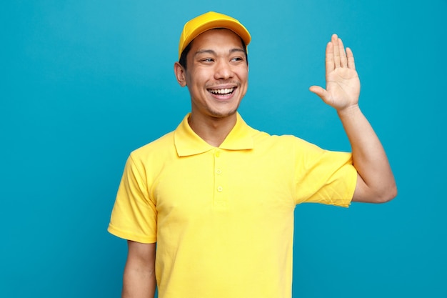 Joyful young delivery man wearing uniform and cap looking at side doing hi gesture 