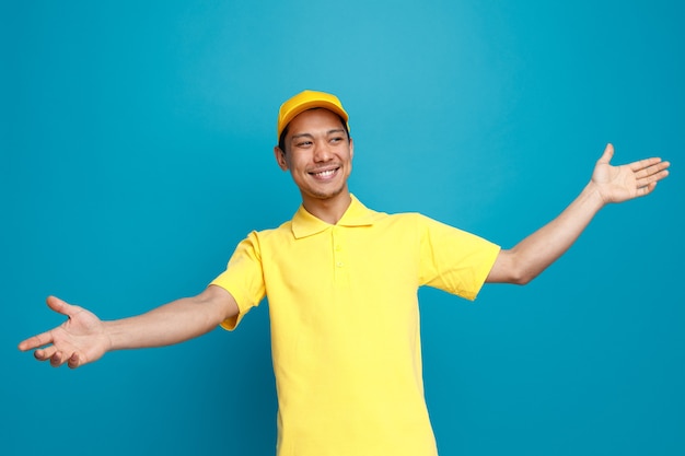 Joyful young delivery man wearing uniform and cap looking at side stretching out hands showing empty hands 