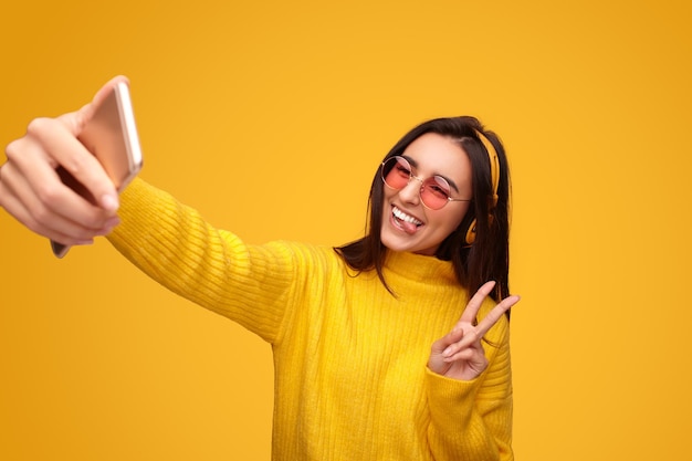 Joyful young ethnic lady smiling and showing peace sign while taking selfie