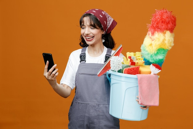 Joyful young female cleaner wearing uniform and bandana holding bucket of cleaning tools and mobile phone looking at mobile phone isolated on orange background