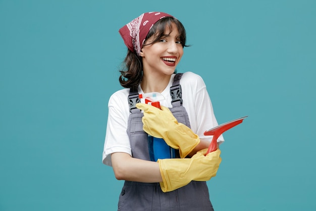 Joyful young female cleaner wearing uniform bandana and rubber gloves holding wiper and cleanser crossed looking at camera isolated on blue background