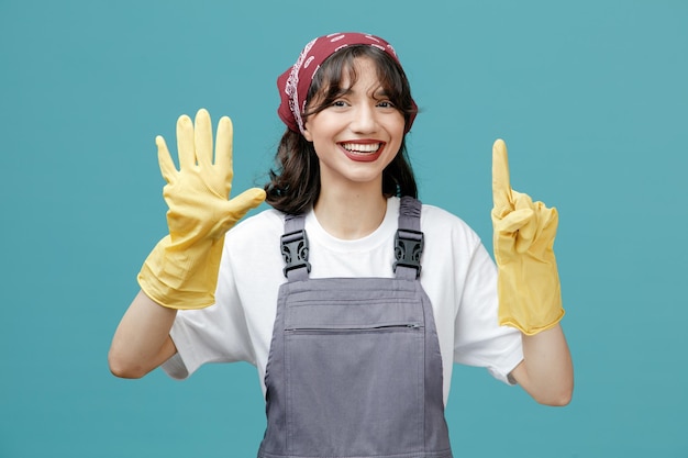 Gioiosa giovane donna addetta alle pulizie che indossa bandana uniforme e guanti di gomma guardando la fotocamera che mostra sei con le mani isolate su sfondo blu