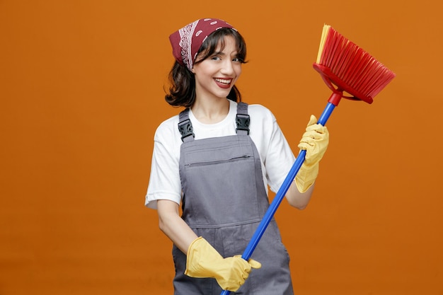 Gioiosa giovane donna addetta alle pulizie che indossa guanti di gomma uniformi e bandana che tiene il mocio del tergipavimento con entrambe le mani guardando la fotocamera isolata su sfondo arancione