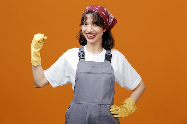 Joyful young female cleaner wearing uniform rubber gloves and bandana showing be strong gesture while keeping hand on waist and looking at camera isolated on orange background