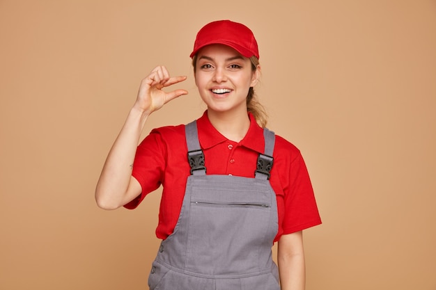 Joyful young female construction worker wearing uniform and cap doing small amount gesture 