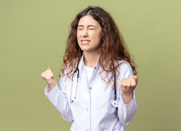 Joyful young female doctor wearing medical robe and stethoscope doing yes gesture with closed eyes isolated on olive green wall