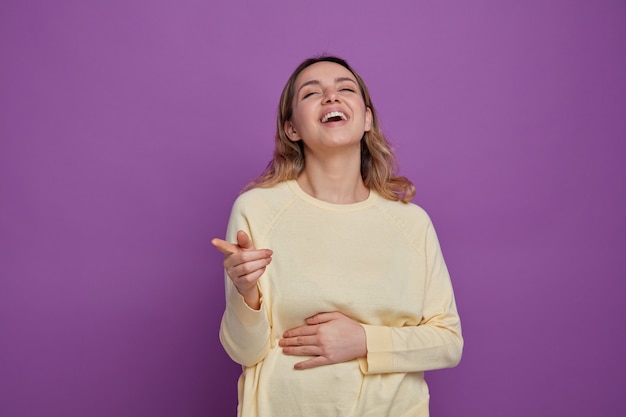 Joyful young girl keeping hand on belly pointing at side laughing 