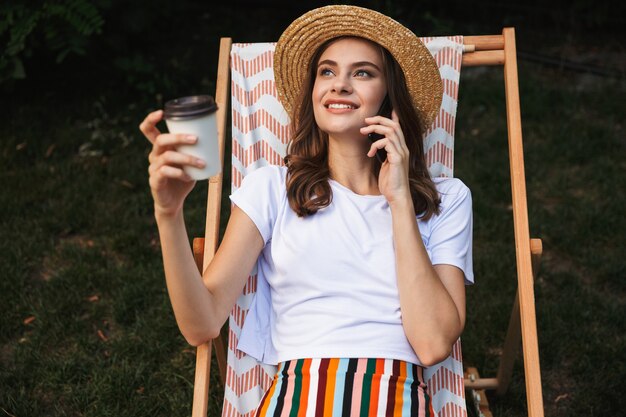 Joyful young girl resting on a hammock at the city park outdoors in summer, drinking takeaway coffee, talking on mobile phone