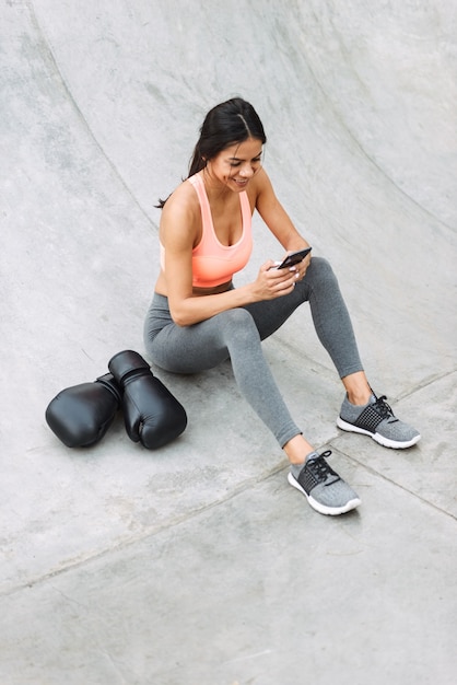 joyful young girl in sportswear holding cellphone while sitting on concrete floor with boxing gloves