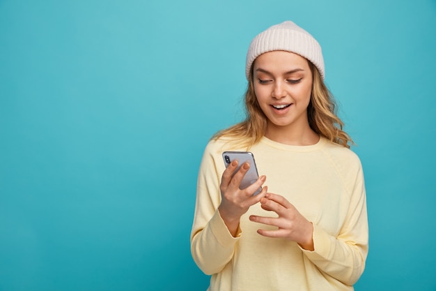 Joyful young girl wearing winter hat holding and looking at mobile phone 