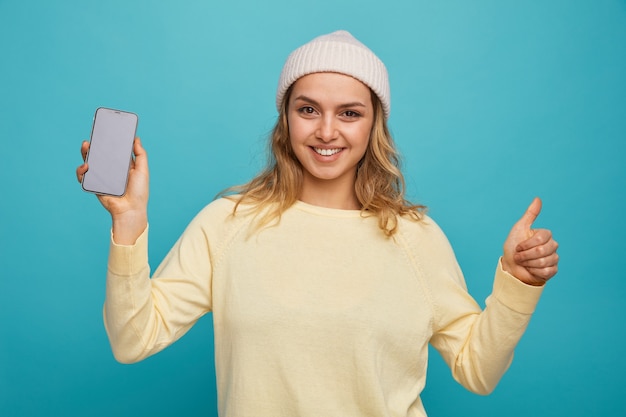 Joyful young girl wearing winter hat holding mobile phone showing thumb up 