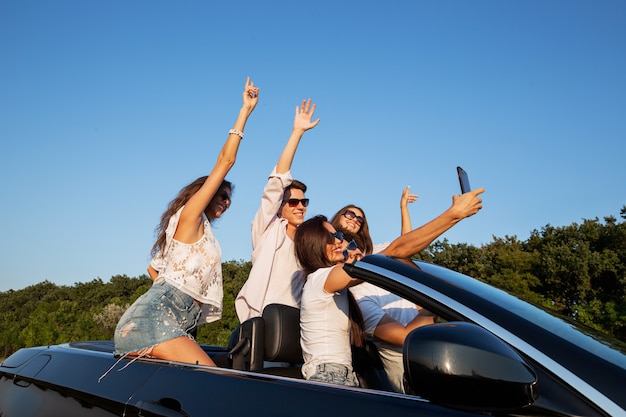 Joyful young girls and guys are sitting in a black cabriolet on the road and making selfie on a sunny day. .