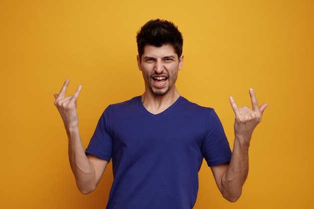 Joyful young handsome man looking at camera doing rock gesture on yellow background