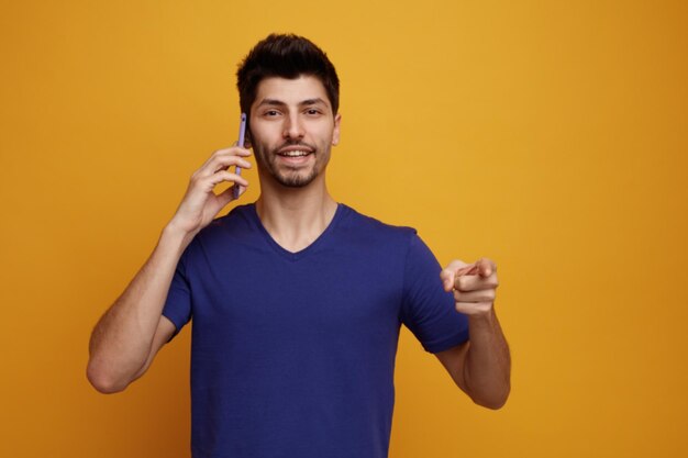 Joyful young handsome man talking on phone looking and pointing at camera on yellow background