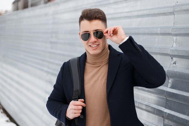 Joyful young hipster man in black sunglasses in a stylish knitted sweater with a black backpack in an elegant coat posing beside a shiny metal fence