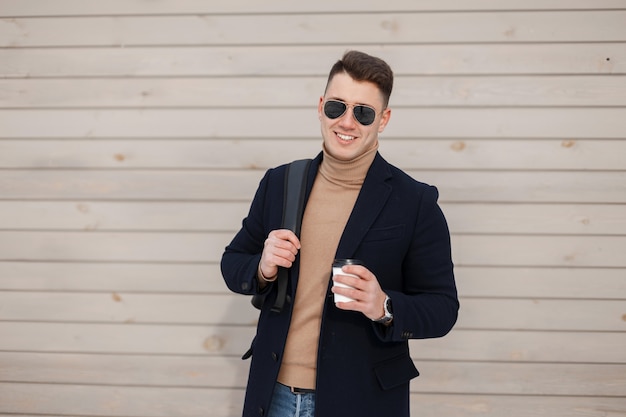 Joyful young hipster man in stylish sunglasses in fashionable spring clothes with a trendy backpack is standing near the wooden wall and holding a cup with coffee outdoors