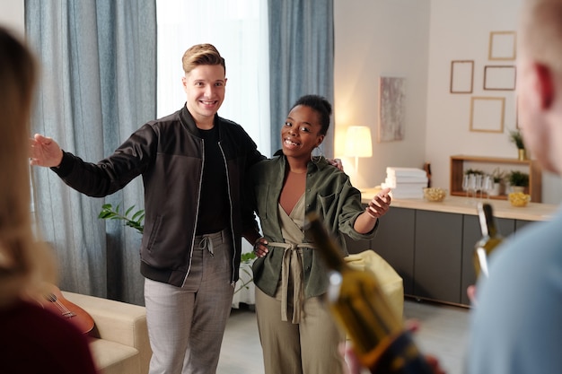 Photo joyful young intercultural couple meeting their friends with bottles of wine while guy talking to blond woman and inviting guests to come in