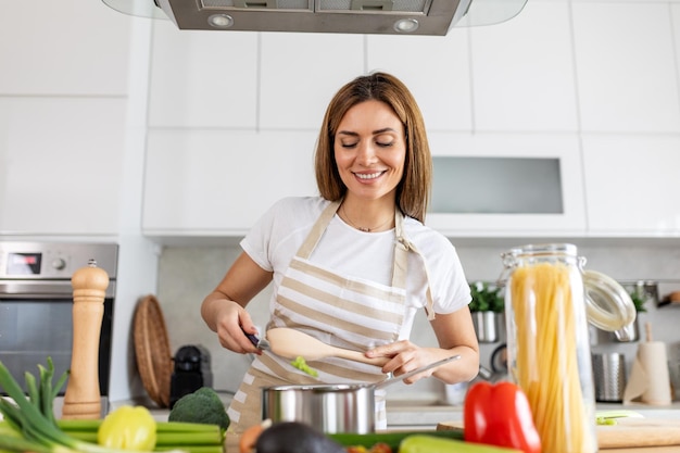 Foto giovane signora gioiosa cucina e assaggia la cena in una pentola presente in una cucina domestica contemporanea casalinga prepara un pasto nutriente con un sorriso vita domestica e nutrimento alimentazione sana