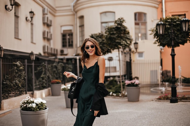 Joyful young lady with brunette wavy hair stylish sunglasses long silk dress and black bag walking on street smiling and posing against city architecture background