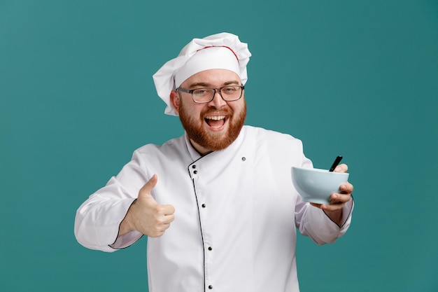 Joyful young male chef wearing glasses uniform and cap showing empty bowl with spoon in it looking at camera showing thumb up isolated on blue background