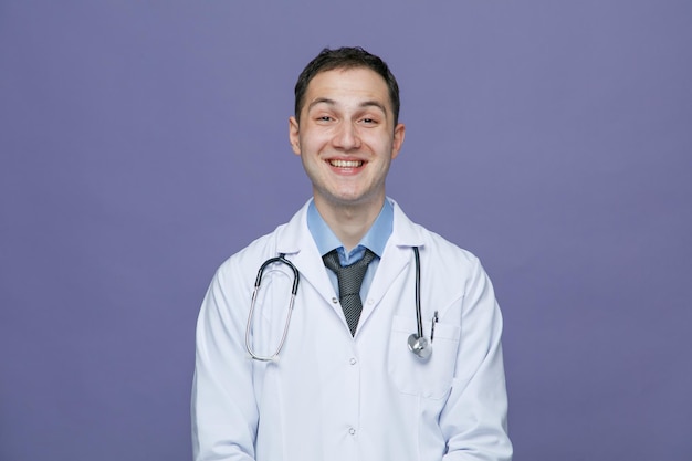 Gioioso giovane medico maschio che indossa un abito medico e uno stetoscopio intorno al collo guardando la fotocamera sorridente isolata su sfondo viola