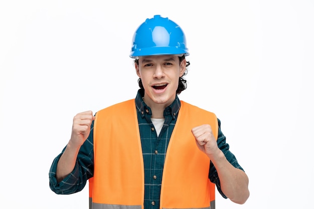 Joyful young male engineer wearing safety helmet and safety vest looking at camera doing yes gesture isolated on white background