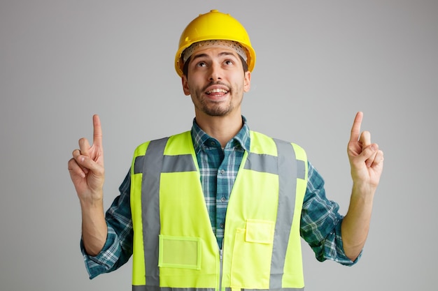 Радостный молодой инженер-мужчина в защитном шлеме и униформе смотрит и указывает вверх на белом фоне