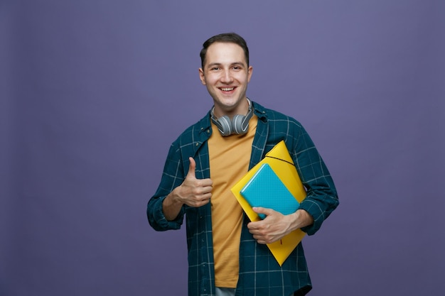 Joyful young male student wearing headphones around neck holding folder and note book looking at camera showing thumb up isolated on purple background