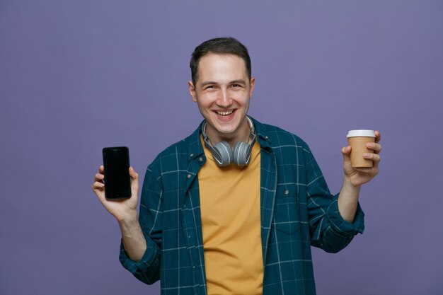 joyful young male student wearing headphones around neck holding paper coffee cup looking at camera showing mobile phone isolated on purple background