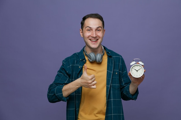 Joyful young male student wearing headphones around neck looking at camera showing alarm clock and thumb up isolated on purple background