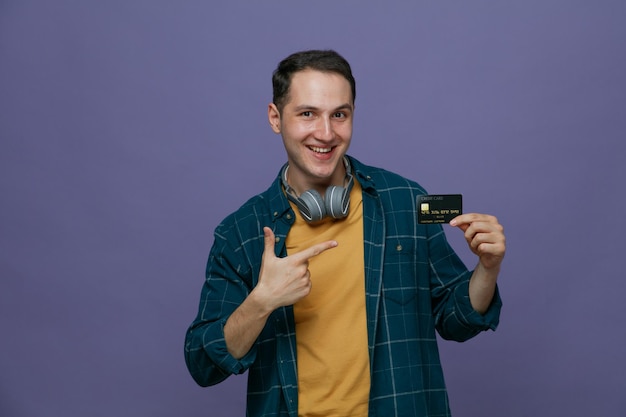 Joyful young male student wearing headphones around neck showing credit card pointing at it looking at camera isolated on purple background