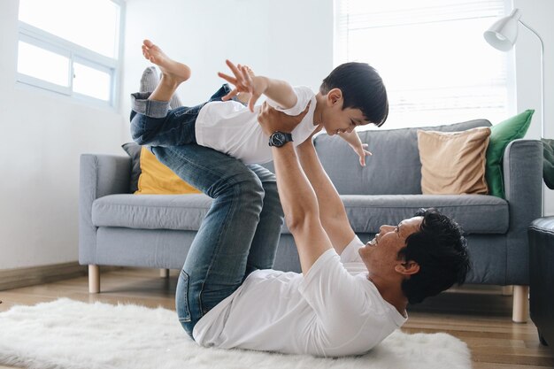 Joyful young man father lying on carpet floor lifting excited happy little child son at home