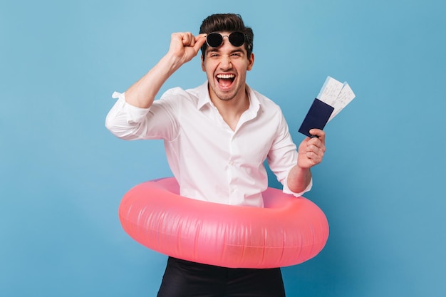 Joyful young man laughs wholeheartedly holding passport tickets
and inflatable circle portrait of man in white shirt taking off
sunglasses