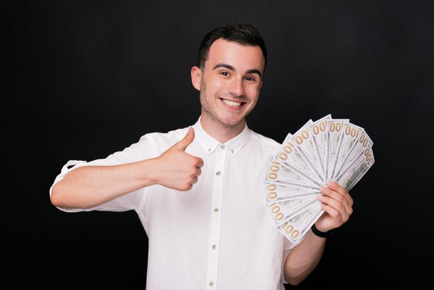 Joyful young man shows thumb up gesture and with another hand holding some dollars