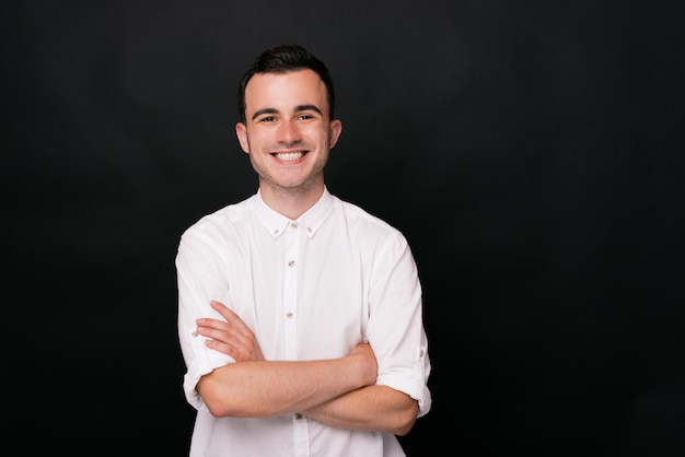 Joyful young man in white shirt is smiling at the camera on black background crossing arms.