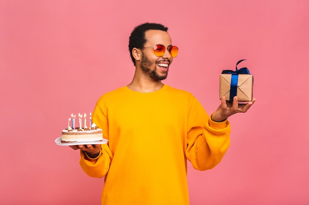 Joyful young man with gift box blowing candles on a birthday cake isolated on pink.