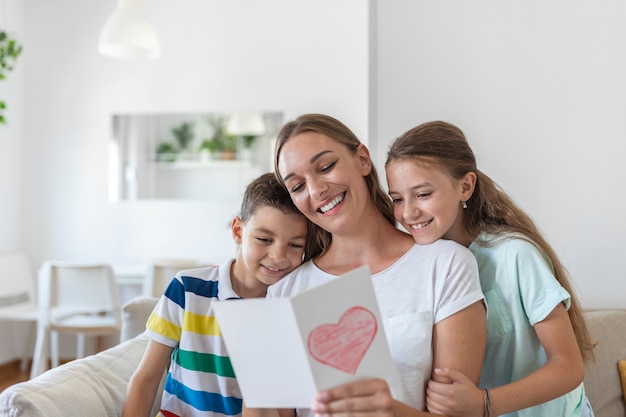 Joyful young mother and little children sitting on sofa and embracing while reading wishes and congratulations with holiday in presented postcard at home