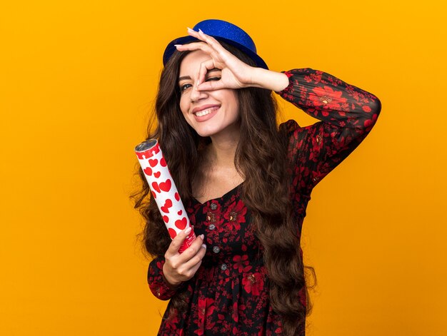 Радостная молодая тусовщица в партийной шляпе держит конфетти-пушку, делая жест взгляда, изолированного на оранжевой стене с копией пространства