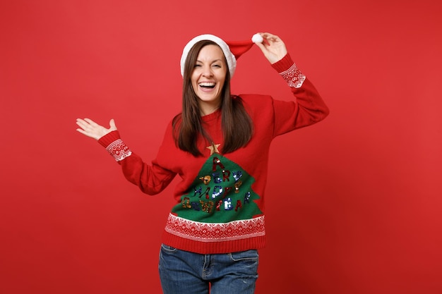 빨간 벽 배경에 격리된 손을 옆으로 가리키는 크리스마스 모자에 손을 얹고 니트 스웨터를 입은 즐거운 어린 산타 소녀. 새해 복 많이 받으세요 2019 축 하 휴일 파티 개념입니다. 복사 공간을 비웃습니다.