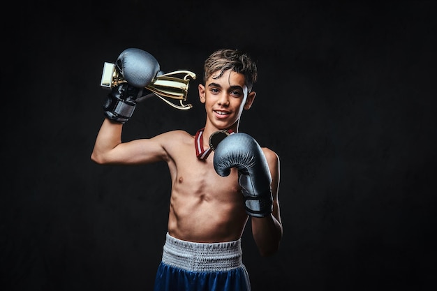 Il gioioso giovane campione di pugili senza maglietta che indossa guanti tiene una coppa del vincitore e la medaglia d'oro. isolato su sfondo scuro.