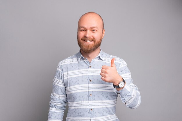 Joyful young smiling bearded man showing thumb up over grey background