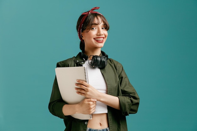 joyful young student girl wearing bandana and headphones around neck holding large note pad with both hands looking at camera isolated on blue background