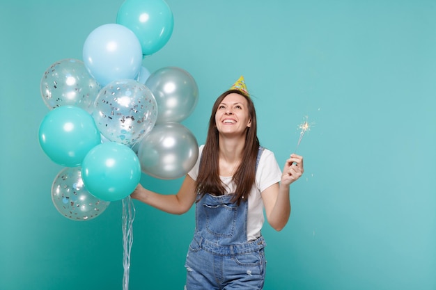 Joyful young woman in birthday hat looking up, holding burning sparkler, celebrating with colorful air balloons isolated on blue turquoise background. Birthday holiday party, people emotions concept.