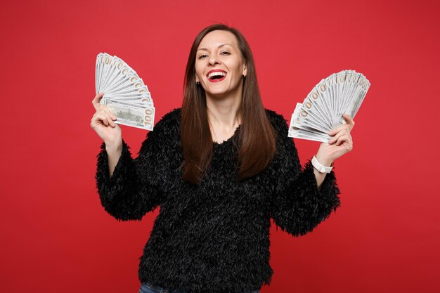 Joyful young woman in black fur sweater holding fan of money in dollar banknotes cash money isolated on bright red background in studio. People sincere emotions, lifestyle concept. Mock up copy space.