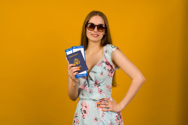 Joyful young woman in blue dress with flowers and sunglasses is holding airline tickets with a passport on a yellow background. rejoices in the resumption of tourism after the coronovirus pandemic