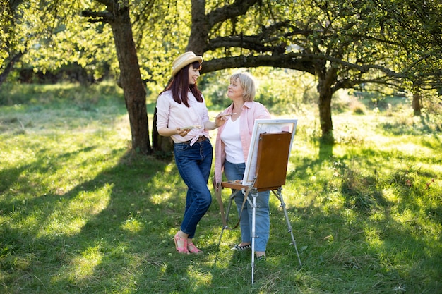 캐주얼웨어와 밀짚 모자에 즐거운 젊은 여자, 그녀의 성숙한 어머니와 함께 그림을 그리는 동안 야외 정원에서 재미. 공동 활동, 창의성 개념.