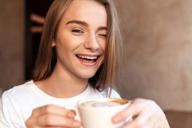 Foto gioiosa giovane donna che beve caffè e ammicca mentre è seduta in un accogliente caffè