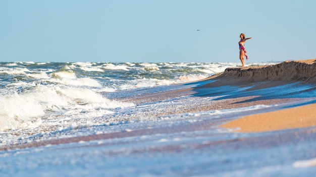 Joyful young woman enjoys stormy waves of the sea looking far beyond the horizon on a sunny summer day while relaxing on the sea. Concept of vacation and privacy. Copyspace