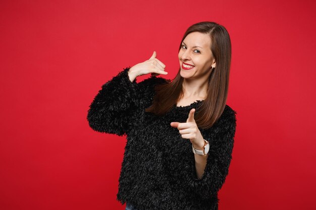 Joyful young woman in fur sweater doing phone gesture like says call me back, pointing index finger on camera isolated on red background. People sincere emotions lifestyle concept. Mock up copy space.
