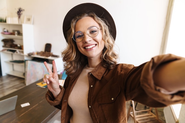 joyful young woman in hat smiling and gesturing peace sign while taking selfie photo indoor